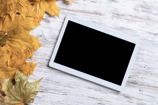 Tablet computer with blank screen lies on vintage wooden desk with bright foliage. Flat lay composition with autumn leaves on white wooden surface. Internet communication and digital technology