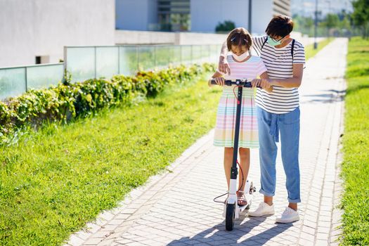 Mother teaching her daughter to ride an electric scooter in urban background wearing surgical masks
