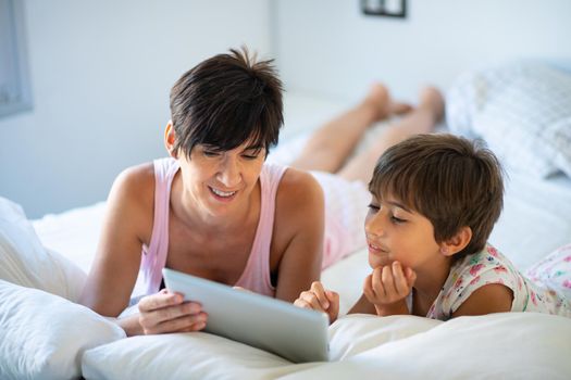 Funny mom and lovely little girl are having fun on the bed. Middle-age mother with her eight years daughter using digital tablet in bedroom.