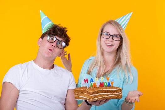 Funny young couple in paper caps and with a cake make a foolish face and wish happy birthday while standing against a yellow background. Concept of congratulations and fooling around