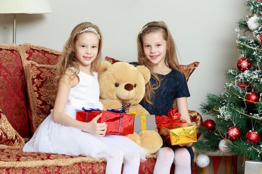 Cute little twin girls, sitting on the floor near the Christmas tree and electric fireplace on which candles are burning. In the hands of the girls boxes with gifts.