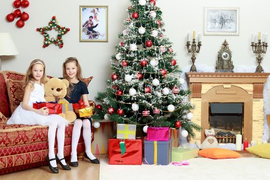 Cute little twin girls, sitting on the floor near the Christmas tree and electric fireplace on which candles are burning. In the hands of the girls boxes with gifts.