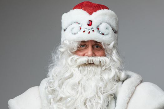 Stock photo of senior Father Frost wearing decorated winter hat and white long beard looking at camera and smiling with his kind eyes. Isolate on grey background.