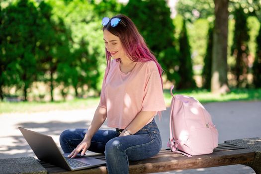 Young girl with pink hair is studying in the spring park, sitting on the wooden bench and browsing on her laptop