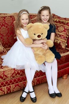 Two adorable little twin girls sitting on sofa hugging a big Teddy bear.