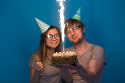 Funny young couple in paper caps and with a cake make a foolish face and wish happy birthday while standing against a blue background. Concept of congratulations and fooling around