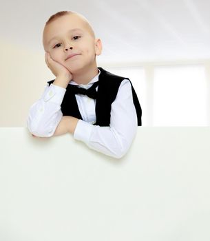 Cute little blonde boy in a black waistcoat and a white shirt and a bow at the neck , peeping over white banner.He put his head on his hand.On a gray background.