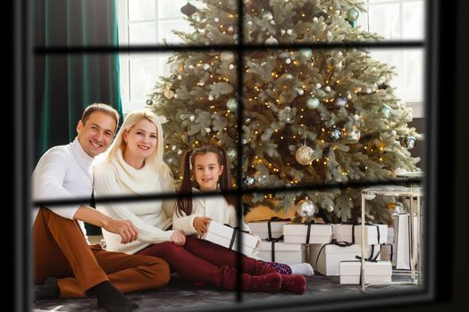 Young big family celebrating Christmas enjoying dinner, view from outside through a window into a decorated living room with tree and candle lights, happy parents eating with three kids