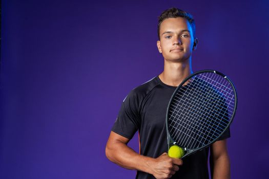 Caucasian young man tennis player posing with tennis racket against purple background close up