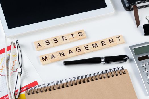 Asset management concept with letters on cubes. Still life of office workplace with supplies. Flat lay white surface with tablet computer, calculator and notepad. Investment strategy and planning.