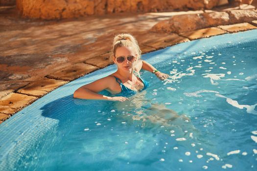 Woman in blue swimsuit and sunglasses relaxing in outdoor pool with clean transparent turquoise water. Woman sunbathing in bikini at tropical resort.