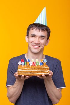 Crazy cheerful young guy with glasses holding a burning candle in his hands and a congratulatory cake on a yellow background. Birthday and anniversary celebration concept. Advertising space