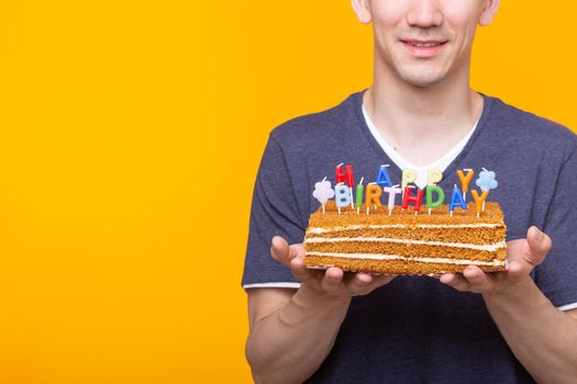 Funny positive guy in glasses holds in his hands a cake with the inscription happy birthday posing on a yellow background. Concept of holidays and anniversaries. Advertising space