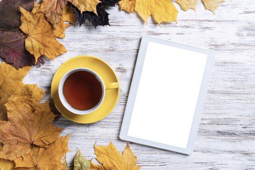 Tablet computer and cup of black tea lies on vintage wooden desk with bright foliage. Flat lay composition with autumn leaves on white wooden surface. Internet communication and digital technology