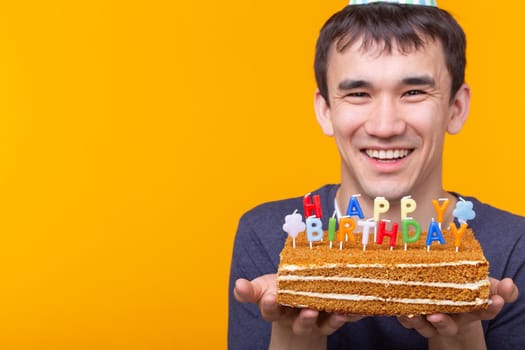 Crazy cheerful young guy with glasses holding a burning candle in his hands and a congratulatory cake on a yellow background. Birthday and anniversary celebration concept. Advertising space