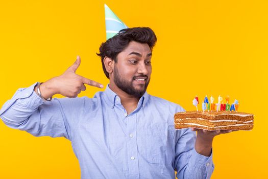 Positive young man holding a happy birthday cake and two burning bengal lights posing on a yellow background. Advertising space