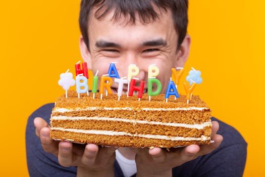 Positive funny young asian guy with a cap and a burning candle and a cake in his hands posing on a yellow background. Anniversary and birthday concept. Advertising space