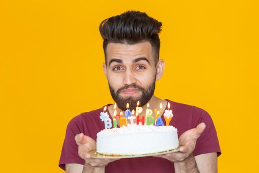Close-up young handsome man blows off a candle from a burning cake posing for a yellow background. Holiday concept. Place for advertising