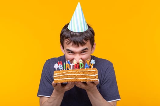 Portrait of a funny positive guy with a paper cap and glasses holding a congratulatory cake in his hands on a yellow background. Concept and fun and celebration. Advertising space