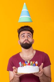 Positive handsome young hipster man in burgundy t-shirt holding congratulatory cake with inscription happy birthday posing on a yellow background. Concept of congratulations and anniversaries