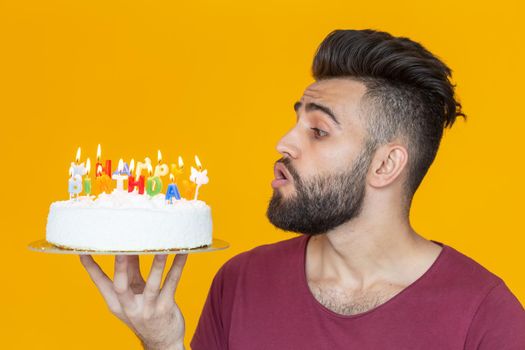 Side view of a handsome young bearded man blowing off candles with a congratulatory cake posing on a yellow background. Advertising space