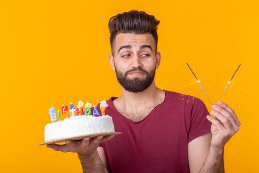 Positive funny young asian guy with a cap and a burning candle and a cake in his hands posing on a yellow background. Anniversary and birthday concept. Advertising space