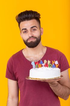 Embarrassed young male hipster with beard holding a birthday cake in his hands and looking thoughtfully at him posing on a yellow background. Concept of time quickly flies by. Advertising space