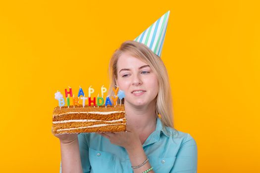 Positive funny young woman with a cap and a homemade cake in her hands posing on a yellow background. Anniversary and birthday concept