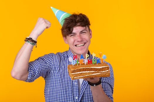 Positive funny young guy with a cap and a burning candle and a cake in his hands posing on a yellow background. Anniversary and birthday concept. Advertising space