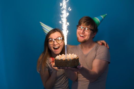 Young cheerful students charming girl and nice guy in greeting paper caps holding a cake with a bengal sparks candle. Concept of congratulations on the holiday and anniversary