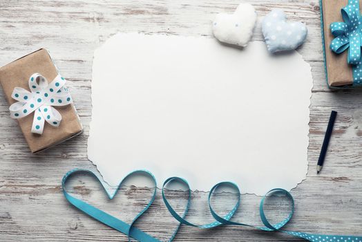 Flat lay composition with blue ribbon in shape of heart. Blank sheet of white paper and gift box on vintage wooden table. Happy birthday or anniversary congratulation. Romantic event invitation.