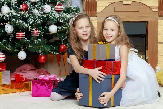 Cute little twin girls, sitting on the floor near the Christmas tree and electric fireplace on which candles are burning. In the hands of the girls boxes with gifts.