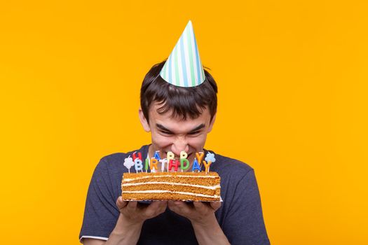 Funny positive guy in glasses holds in his hands a cake with the inscription happy birthday posing on a yellow background. Concept of holidays and anniversaries. Advertising space