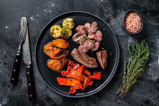 Roasted lamb or goat tenderloin Meat steak in plate with grilled vegetables. Black wooden background. Top view.
