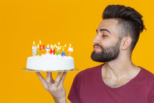 Side view of a handsome young bearded man blowing off candles with a congratulatory cake posing on a yellow background. Advertising space