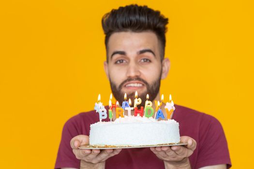 Close-up young handsome man blows off a candle from a burning cake posing for a yellow background. Holiday concept. Place for advertising