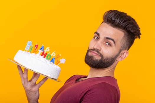 Embarrassed young male hipster with beard holding a birthday cake in his hands and looking thoughtfully at him posing on a yellow background. Concept of time quickly flies by. Advertising space