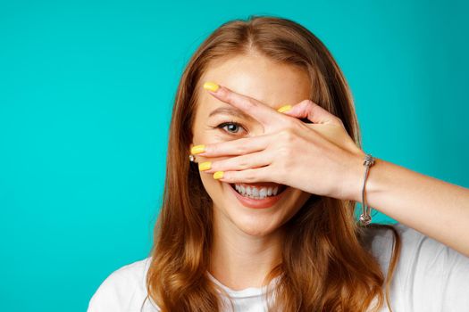 Happy young woman peeping through her fingers and smiling against blue background