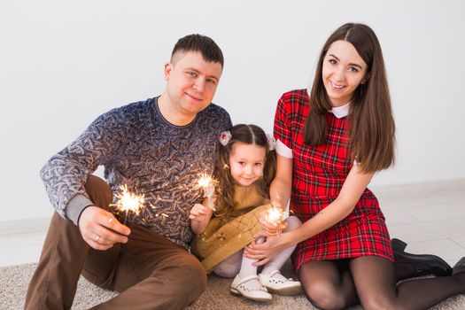 Celebration, family and holidays concept - happy parents and little daughter with sparklers sitting on carpet over grey background.