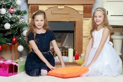 Cute little twin girls, sitting on the floor near the Christmas tree and electric fireplace on which candles are burning.