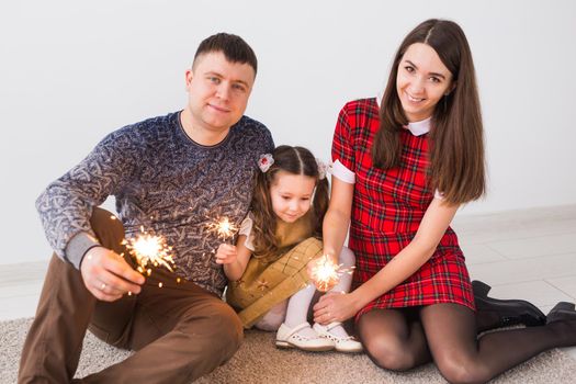 Celebration, family and holidays concept - happy parents and little daughter with sparklers sitting on carpet over grey background.