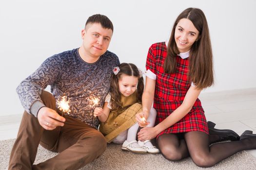 Celebration, family and holidays concept - happy parents and little daughter with sparklers sitting on carpet over grey background.