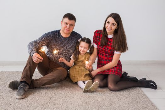 Celebration, family and holidays concept - happy parents and little daughter with sparklers sitting on carpet over grey background.