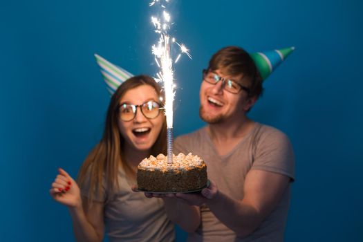 Young cheerful students charming girl and nice guy in greeting paper caps holding a cake with a bengal sparks candle. Concept of congratulations on the holiday and anniversary