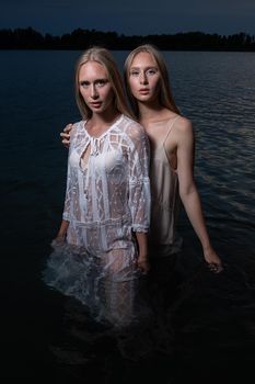 two young twin sisters with long blond hair posing in light dresses in water of lake at summer night, looking at camera. stylish fashion photoshoot with flashlight. outdoors evening photosession