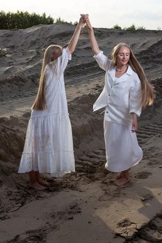 two young blond pretty twins dancing at sand quarry in elegant white dress, skirt, jacket. stylish fashion photoshoot, summer photosession. identical sisters spend time together outdoors, have fun