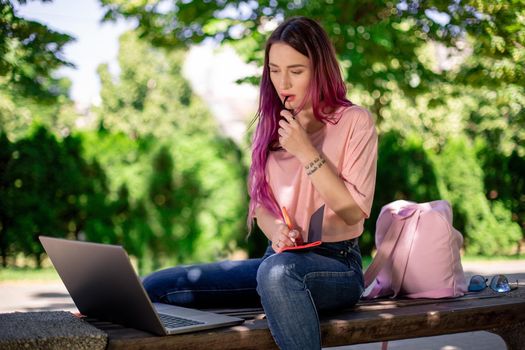 Woman writing in a notebook sitting on a wooden bench in the park. Girl working outdoors on portable computer, copy space. Technology, communication, freelance and remote working concept.