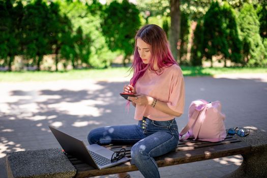 Woman writing in a notebook sitting on a wooden bench in the park. Girl working outdoors on portable computer, copy space. Technology, communication, freelance and remote working concept.