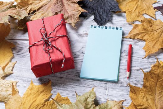 Spiral notepad with pen and gift box lies on vintage wooden desk with bright foliage. Flat lay composition with autumn leaves on white wooden surface. Happy thanksgiving holiday congratulation.