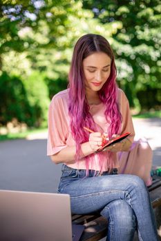 Woman writing in a notebook sitting on a wooden bench in the park. Girl working outdoors on portable computer, copy space. Technology, communication, freelance and remote working concept.